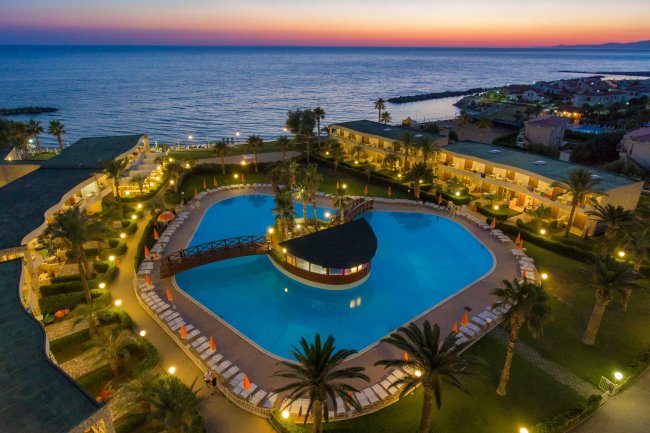 Vista dall'alto di un resort al tramonto, con una grande piscina centrale e giardino.