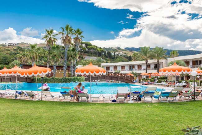 Piscina all'aperto con ombrelloni arancioni e lettini, circondata da palme e colline verdi.