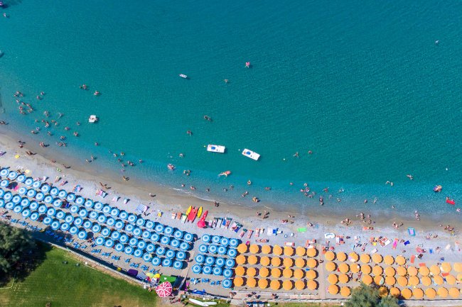 Spiaggia vista dall'alto, con ombrelloni blu e arancioni e mare azzurro.