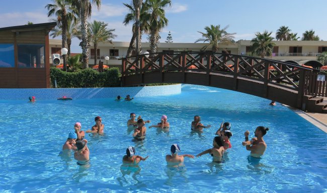 Persone svolgono esercizi in piscina, ponte di legno e palme.