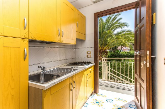Cucina con mobili gialli, lavello e porta aperta verso un balcone con palme.