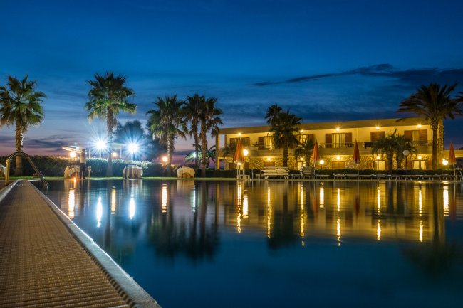 Piscina illuminata di notte con palme e un edificio sullo sfondo.