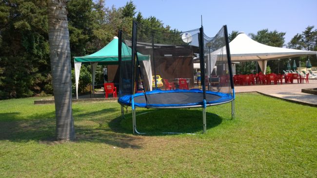 Un prato con trampolino blu e tende con tavoli e sedie rosse.