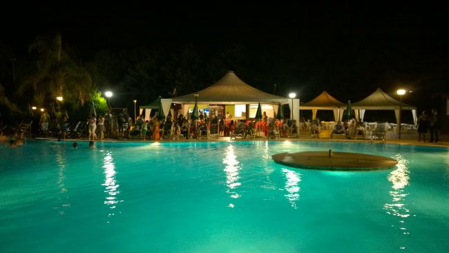 Piscina illuminata di notte con persone sedute sotto gazebo.
