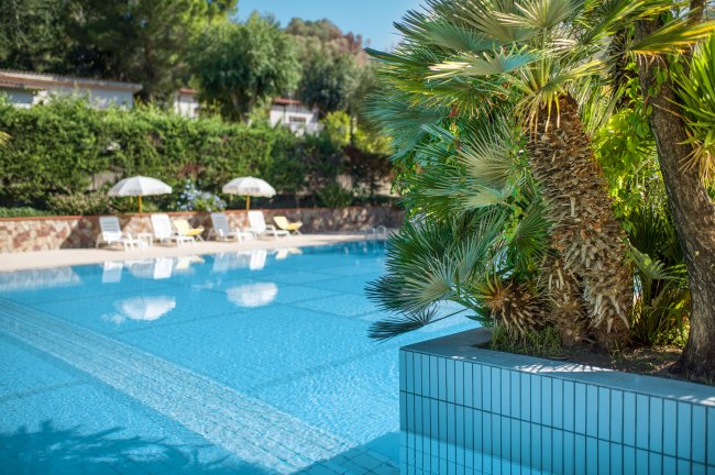 Lato di una piscina con piante rigogliose e lettini con ombrelloni sullo sfondo.