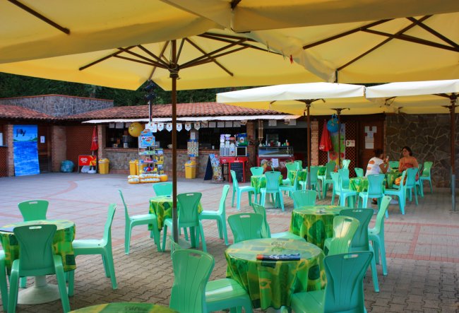Tavolini verdi sotto ombrelloni gialli in uno spazio esterno con bar sullo sfondo.
