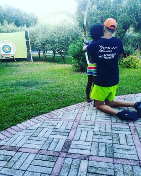 Persona aiuta un bambino a tirare con l'arco verso un bersaglio su un prato.