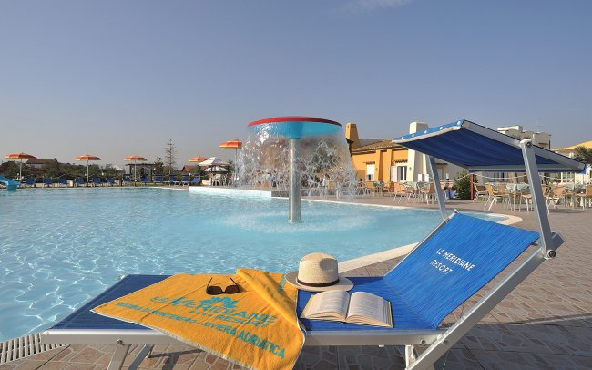 Lettino con asciugamano e cappello vicino a una piscina con fontana, sotto un cielo limpido.
