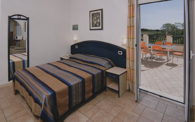 Camera con letto, copriletto a righe blu e marroni, terrazza con tavolo e sedie arancioni.