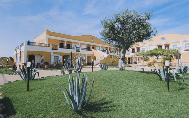 Edificio giallo con balconi, prato verde e piante di agave sotto un cielo azzurro.