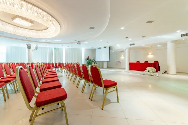 Sala conferenze con sedie rosse, podio e schermo proiettore.