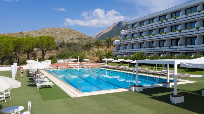 Piscina con lettini e ombrelloni, un hotel sullo sfondo e montagne in lontananza.