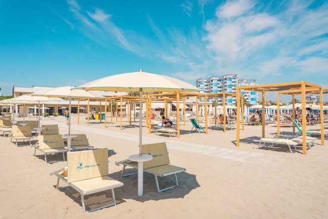 Spiaggia con lettini e ombrelloni bianchi, cielo azzurro e palazzi in lontananza.