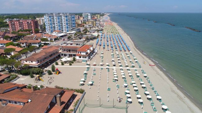 Ampia spiaggia con file di ombrelloni bianchi e verdi, edifici alti sul lungomare.