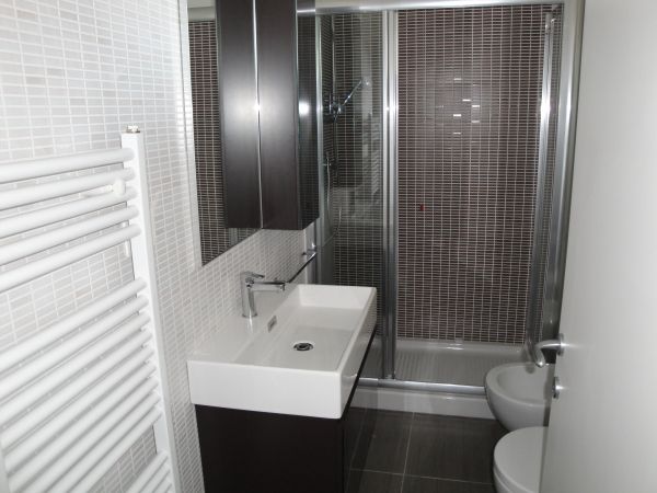Bagno con doccia, lavabo rettangolare bianco, piastrelle marroni, e un termosifone verticale.