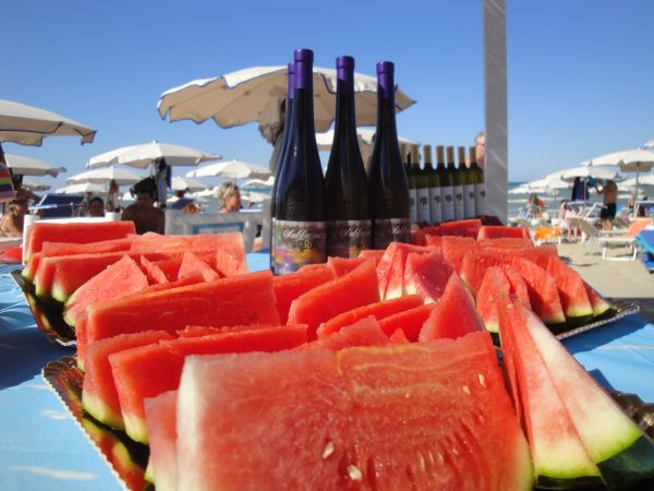 Fette di anguria e bottiglie di vino su un tavolo sotto ombrelloni da spiaggia.