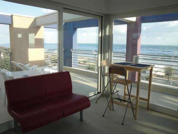 Sala con divano rosso, tavolo alto e vista mare attraverso grandi finestre.