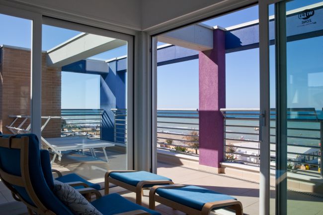 Terrazza vista mare con poltrone blu, tavolini e struttura con elementi colorati.