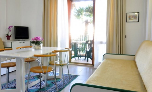 Soggiorno con tavolo, sedie gialle, divano beige e uscita verso un balcone.