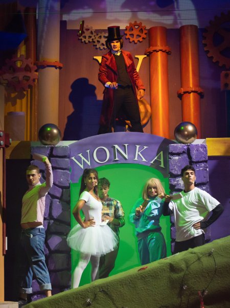 Scena teatrale con personaggi davanti a un ingresso Wonka colorato.