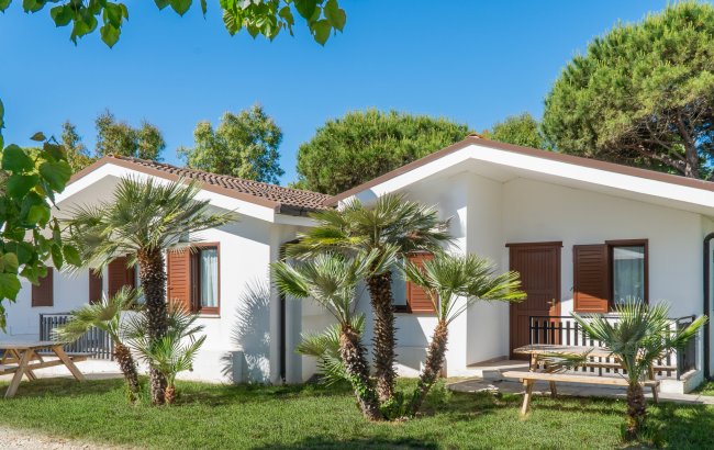 Bungalow bianchi con persiane marroni circondati da alberi e palme sotto un cielo blu.