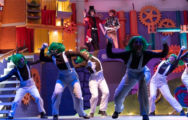 Performers con parrucche verdi ballano su un palco colorato, con due attori sullo sfondo.