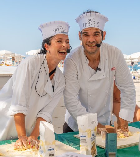 Due chef sorridenti impastano su un tavolo esterno, indossando cappelli da cuoco.