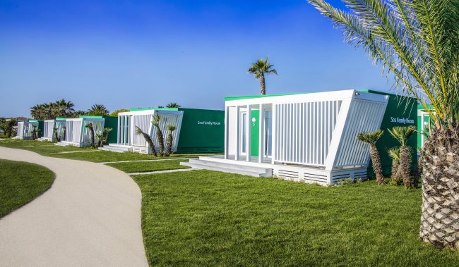 Case modulari bianche e verdi su prato verde con palme e cielo blu.