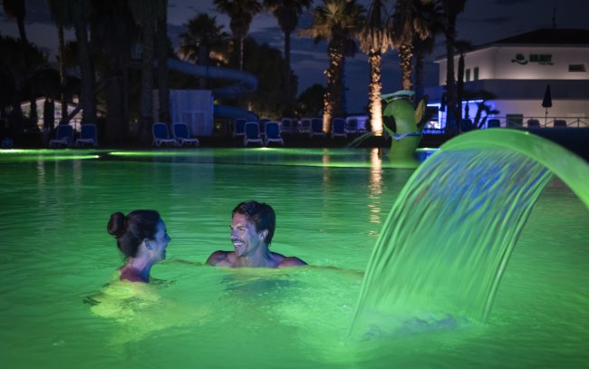 Due persone sorridenti in una piscina illuminata di verde con cascata d'acqua.