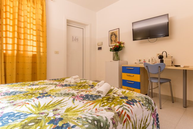 Interno di camera con letto matrimoniale, TV, scrivania con sedia blu e tenda arancione.