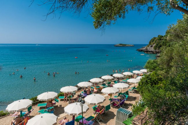 Spiaggia con ombrelloni bianchi e lettini, bagnanti in mare e una vista su una piccola isola.