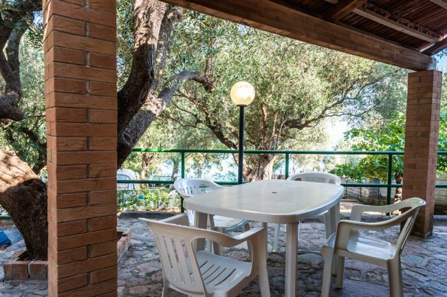 Tavolo bianco con sedie sotto un portico circondato da alberi.