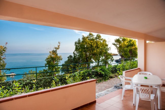Una veranda con tavolo e sedie bianche, vista sul mare e vegetazione.