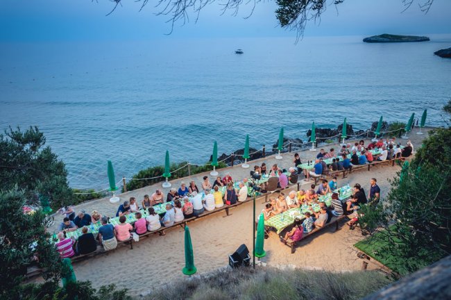 Cena all'aperto accanto al mare, con tavoli pieni di persone e ombrelloni verdi.