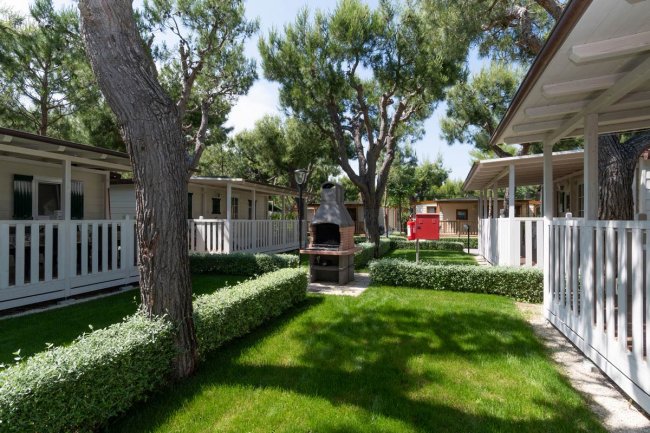 Giardino con barbecue in pietra tra bungalow circondati da siepi e alberi.