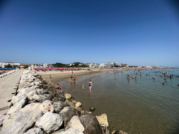 Mare con bagnanti e un lungo molo di rocce. Spiaggia sullo sfondo.