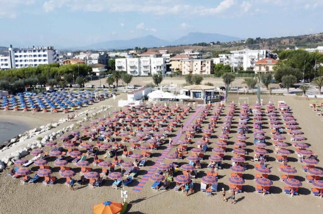 Spiaggia con file ordinate di ombrelloni colorati e lettini, affiancata da palazzi e vegetazione.
