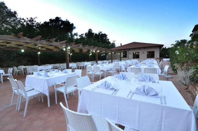 Tavoli all'aperto apparecchiati con tovaglie bianche e sedie su una terrazza, sotto una pergola.