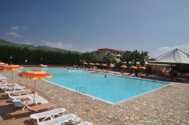 Piscina all'aperto con lettini e ombrelloni colorati. Persone in acqua e cielo sereno.