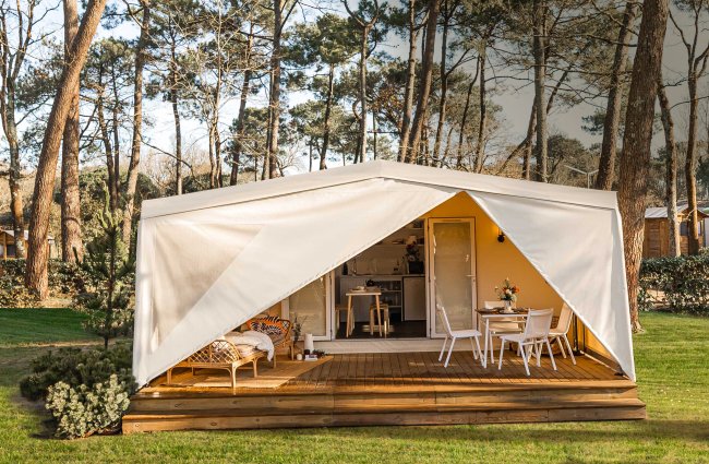Tenda glamping con portico in legno, sedie e tavolo, immersa in un bosco.