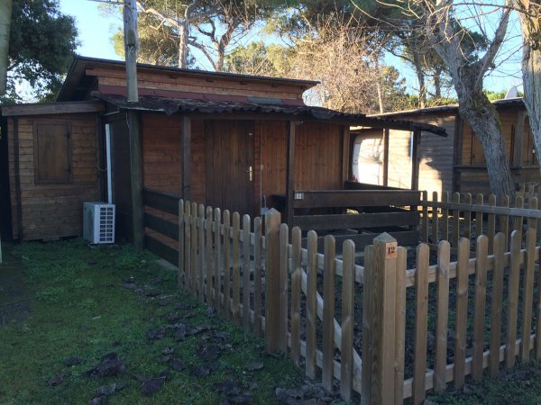 Piccola casa di legno recintata con alberi intorno, porta marrone e aria condizionata esterna.