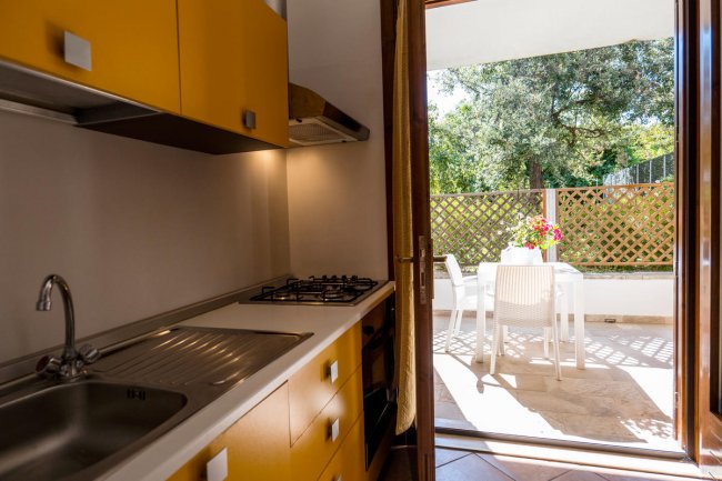Cucina con mobili gialli, lavello, piano cottura e vista su un balcone con tavolo e sedie bianche.