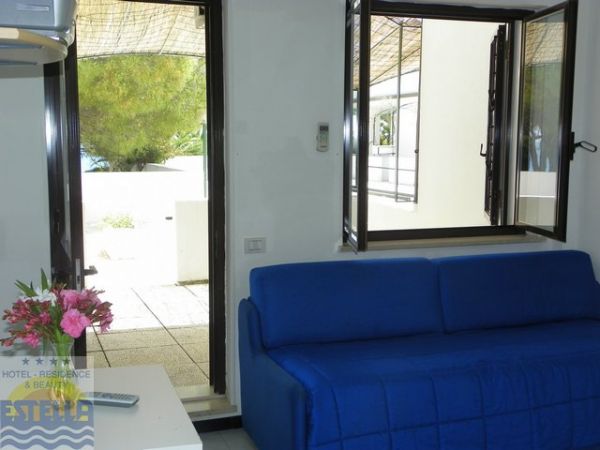 Soggiorno con divano blu, finestra aperta e porta sul terrazzo. Fiori su un tavolo.