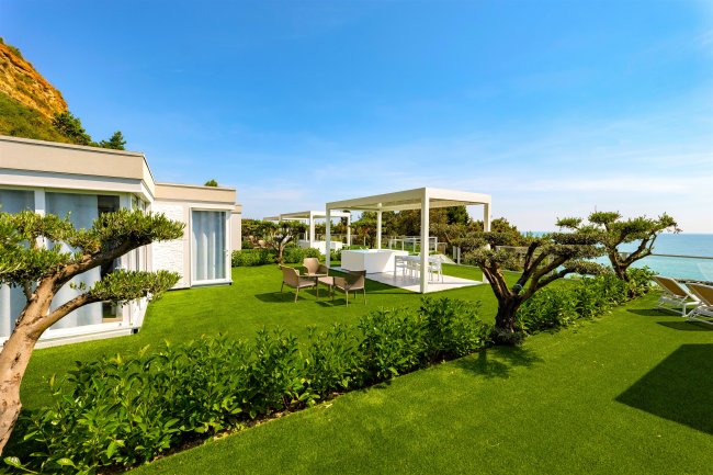 Giardino con prato verde, pergola bianca e vista mare, cielo sereno.