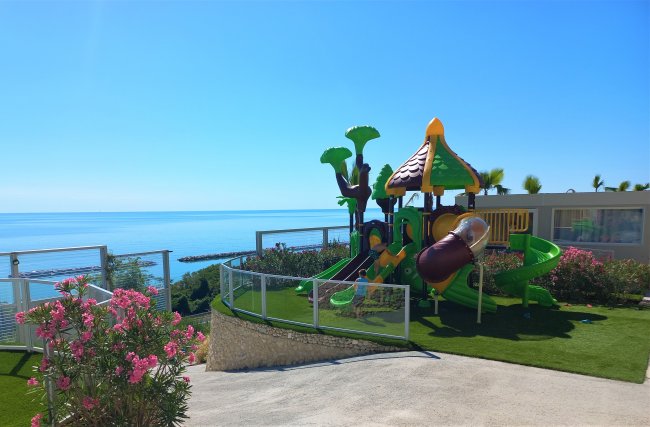 Parco giochi colorato con scivoli, vicino al mare e circondato da aiuole di fiori rosa.