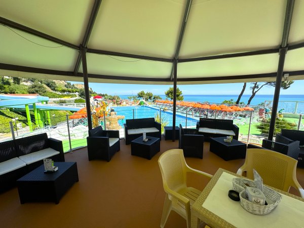 Area coperta con divani e tavolini, vista sul mare e una piscina con lettini arancioni sullo sfondo.