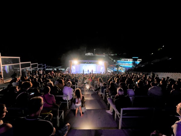 Platea notturna con pubblico seduto e palco illuminato sullo sfondo.