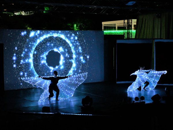 Due performer danzano sul palco con costumi a LED, sfondo di proiezioni luminose.