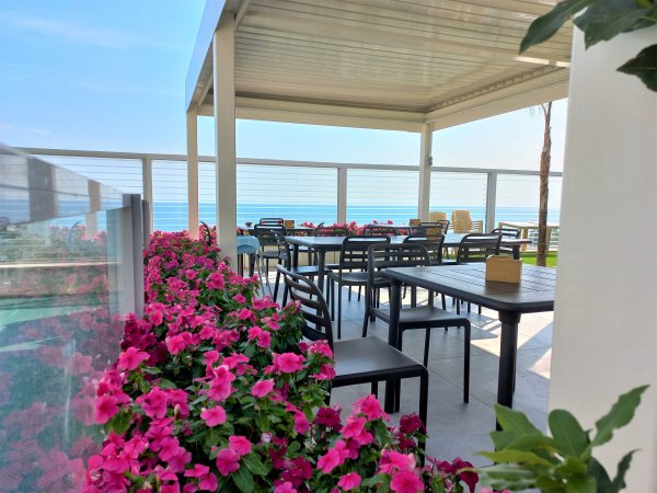 Terrazza con tavoli e sedie, decorata con fiori rosa vivaci, vista sul mare in lontananza.