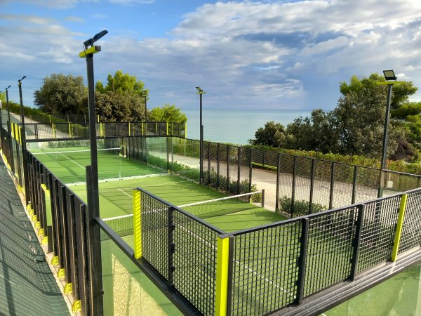 Un campo da padel verde circondato da recinzioni e alberi con vista sul mare.